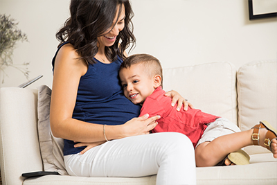 Pregnant woman in blue top sitting on a couch, holding a child in a red shirt who is hugging her.